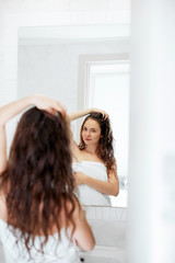 Naklejka premium Hair and body care.Beautiful young woman touching her wet hair with hands and smiling while standing in front of the mirror in bathroom. Girl uses protection moisturizing cream.