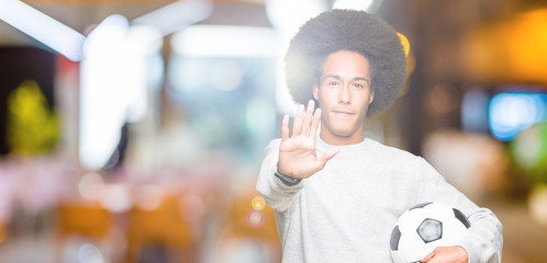 Young african american man with afro hair holding soccer football ball with open hand doing stop...