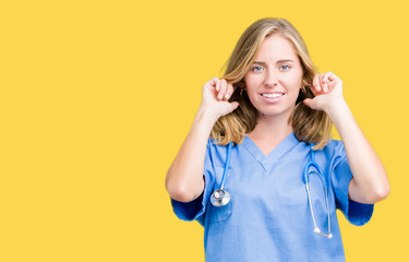 Beautiful young doctor woman wearing medical uniform over isolated background covering ears with fingers with annoyed expression for the noise of loud music. Deaf concept.