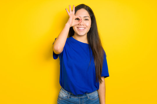 Beautiful brunette woman wearing blue t-shirt over yellow isolated background doing ok gesture with hand smiling, eye looking through fingers with happy face.