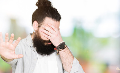 Young man with long hair, beard and earrings covering eyes with hands and doing stop gesture with sad and fear expression. Embarrassed and negative concept.