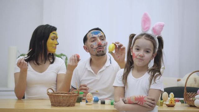 Cute couple is colorizing each other with a help of Easter paint-brush. Their daughter says: "Shame on you I have grown up, but neither you ". Curious situation. Daddy is showing his tongue, mommy is