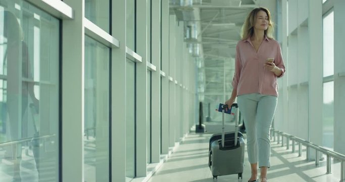 Stylish Caucasian pretty woman going through the airport corridor, carrying suitcase with wheels and typing a message on the smartphone.