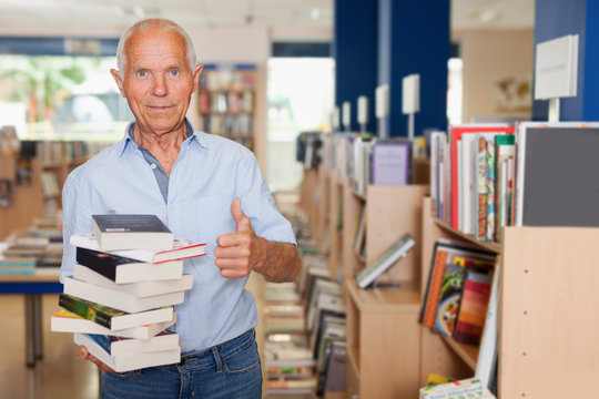 Positive senior man with stack of books in hands in library