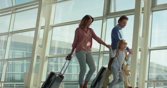 Cheerful Caucasian cute small son with backpack walking the airport hall hand in hand with his parents at both sides with suitcases on wheels while family travelling.