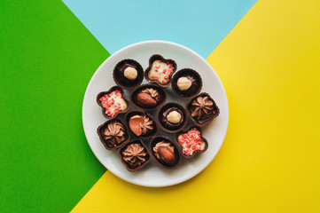 Beautiful chocolates of different shapes and fillings lie in a white plate against the background of colored paper. Yellow, green, blue color.