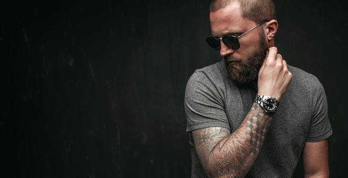 Portrait of a handsome balded man with long well trimmed beard wearing sunglasses and grey shirt looking away to side.