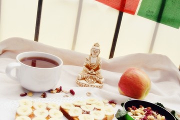 Minimalistic breakfast with dry fruits, cup of tea, apple and decorated bread with banana. Buddha`s statue on background