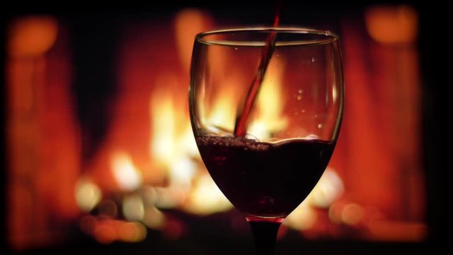 Closeup shoot of red wine being poured into the luxurious empty glass with cozy warm fireplace on the background indoors in a dark apartment