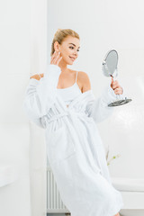 attractive and smiling woman in white bathrobe looking at mirror in bathroom