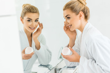 selective focus of attractive and blonde woman in white bathrobe applying face cream and looking at mirror