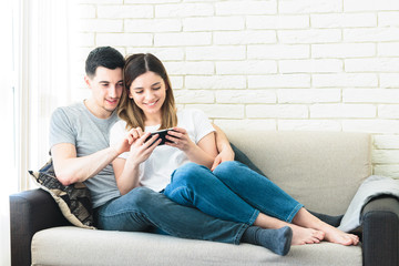 Relaxed couple using a generic mobile phone together sitting on a sofa in the living room at home