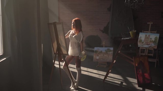 Seductive young red-haired girl is standing in her workroom in only white transparent shirt and painting. The room is in a loft style.