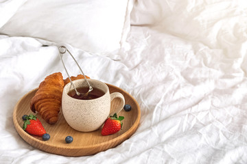 A cup of tea and croissants on the wooden tray standing on the bed with white bedsheets.