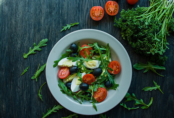 Greek salad with fresh tomatoes, arugula, eggs, olives with olive oil on a dark wood background. Healthy food. Veggie dish.
