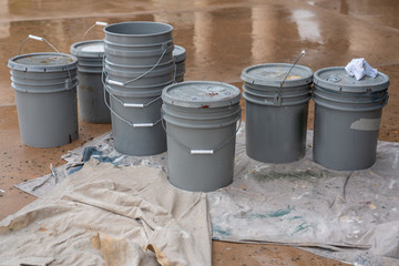 stacks of grey industrial sized paint buckets and a splattered dropcloth outside on a wet pavement