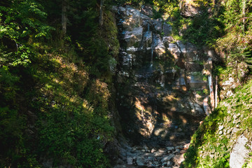 Obraz premium Forest stream going down high rocky waterfall located in between cliffs covered with summer green grass and trees in mountain forest