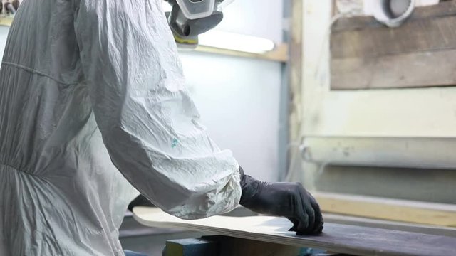 profession, people, carpentry, emotion and people concept -Man paints skateboard in workshop, applies varnish. manufacture of skateboards. he has protective clothing and mask. 4k