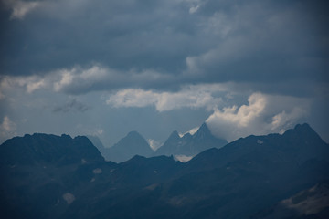 Fototapeta premium The peaks of the Caucasus Mountains in cloudy clouds and the rays of the sun through them in the outskirts of Dombai. And on the tops of snow lies.