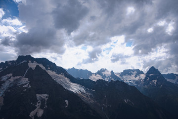 The peaks of the Caucasus Mountains in cloudy clouds and the rays of the sun through them in the outskirts of Dombai. And on the tops of snow lies.