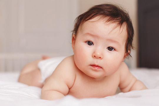 Portrait of cute adorable Asian mixed race baby girl four months old lying on tummy on bed in bedroom looking in camera. Natural emotion face expression. Childhood diversity lifestyle