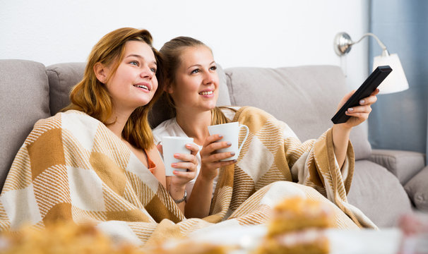 Two young women in plaide are watching TV and warming with tea