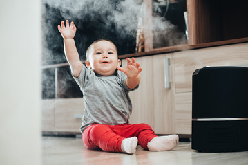 a little girl in red pants looks and touches the humidifier. Moisture in the house concept - obrazy, fototapety, plakaty
