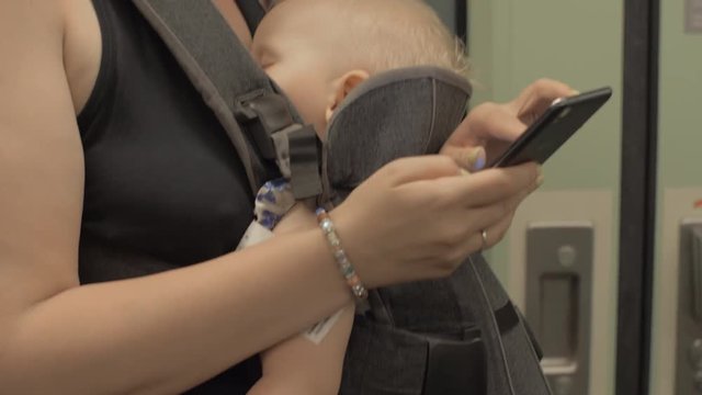 Woman with sleeping baby daughter in carrier riding in subway train and using smartphone