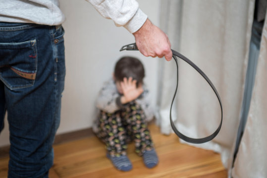 Family violence and aggression concept - furious angry man uses a belt for punishment scared or terrified child boy sitting at wall corner