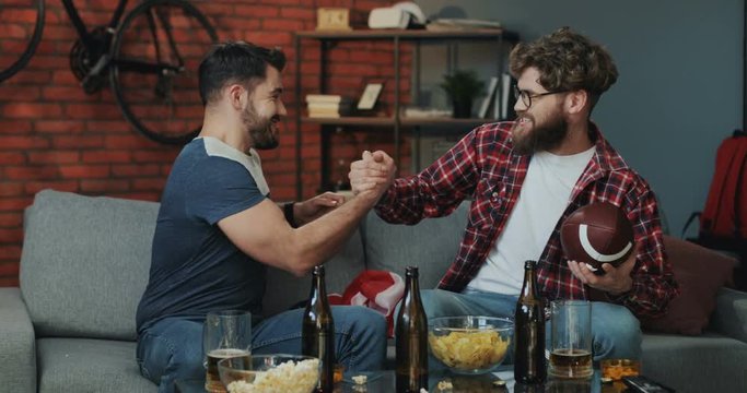 Young Caucasian handsome two men sitting on the couch and watching sport game on TV, then cheering and hugging as celebrating victory of their team in American football match.