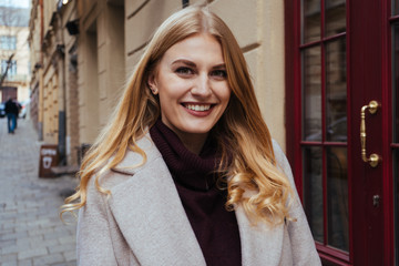 Beautiful blonde woman in stylish coat smiling sincerely at the camera while standing on the city street. Close up portrait shot