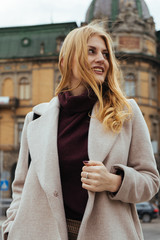 Young pretty woman with blonde hair in casual coat smiling and looking to the side while standing...