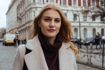 Close up portrait of young pretty woman with blonde hair looking at the camera while standing on the city street on spring day.