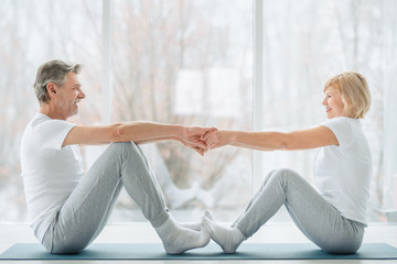 Sports and healthy lifestyle.Sporty middle aged couple sitting on the mat in the white fitness gym and doing stretching while looking each other and smiling. Healthy joints and back. Support trainer.