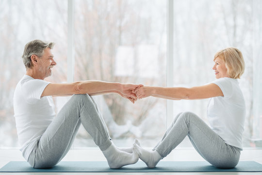 Sports And Healthy Lifestyle.Sporty Middle Aged Couple Sitting On The Mat In The White Fitness Gym And Doing Stretching While Looking Each Other And Smiling. Healthy Joints And Back. Support Trainer.