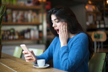 Side view of casual girl watching smartphone and looking amazed with news while sitting in cafeteria