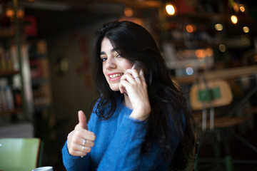 Happy confident beautiful woman gesturing thumbs up looking at camera sitting in a cafe while calling by phone