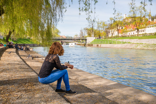 Fototapeta Young woman is sittng on the banks of Ljubljanica river at Trnovo "beach" admiring the beautiful Ljubljana old town in Ljubljana, Slovenia.