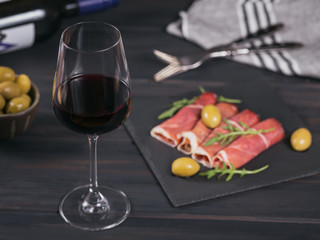 Glass of red wine with slices of cured ham or Spanish jamon serrano or Italian prosciutto crudo, green olives and arugula