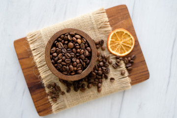 Closeup of coffee beans in wooden rustic bowl on bright background decorated with orange slice.