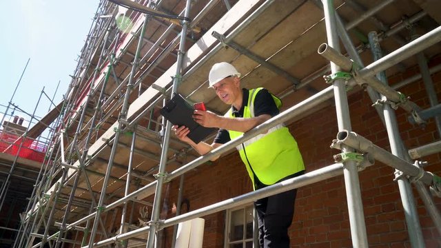 Male builder foreman, worker, surveyor, or architect working on construction building site standing on scaffolding writing on clipboard 