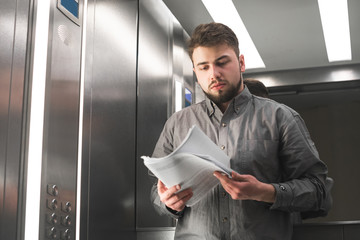Office worker in a shirt standing in an elevator holds documents and reads in his hands. Business man is studying paper in the elevator before work.