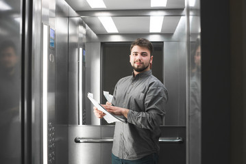 Happy business man in a shirt stands in the elevator with documents in his hands, looks at the camera and smiles. Portrait of a smiling office worker in an elevator, holding paper in his hands © bodnarphoto
