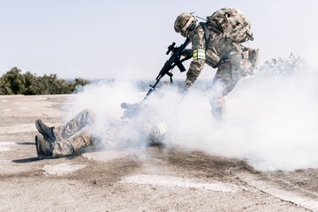 Armed man in camouflage with a sniper gun in his hand, pulling a wounded soldier in the smoke