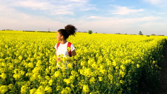 Beautiful happy mixed race African American girl teenager female young woman hiking with red backpack on path through field of yellow flowers. Stabilised tracking shot.