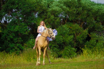 Young rider woman blonde with long hair in a white dress with a train posing and jumping on a...