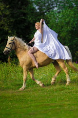 Young rider woman blonde with long hair in a white dress with a train posing and jumping on a...