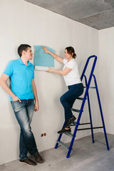 Fototapeta na wymiar Funny couple, woman and her husband wallpapering the walls using a metal ladder. Home renovation and moving to new house concept