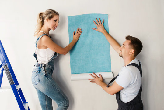 Funny couple, woman and her husband wallpapering the walls using a metal ladder. Home renovation and moving to new house concept