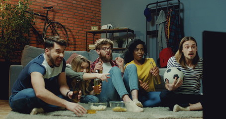 Young mixed-races men and women sitting on the floor at home and watching sport game on TV, then cheering and drinking beer with cheers gesture as celebrating victory.